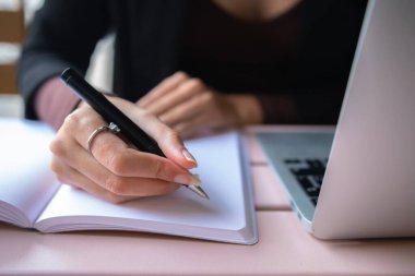 Soft focus of anonymous female freelancer writing down data into planner near netbook while sitting at table and working remotely in cafe