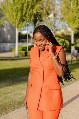 Content modern African American female in vivid orange vest suit and with Afro braids having call on mobile phone walking in sunlight