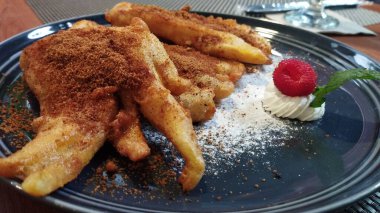 Pisang Goreng Gula Are veya Palm Sugar Muz kızartması pudralanmış palmiye şekeriyle servis edilir artı ahşap bir masada siyah bir tabakta kırmızı böğürtlen garnitürü.