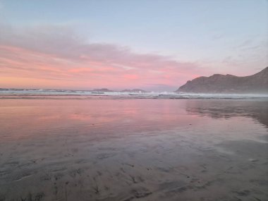 Famara Sahili 'nde gün batımının nefes kesici güzelliğini tecrübe et, Lanzarote. Güneş ufkun altına batarken, gökyüzü sıcak renklerden oluşan bir tuvale dönüşür, engebeli uçurumlara ve sakin Atlantik sularına altın bir parıltı saçar..