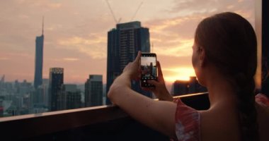 Turist kadın, Malezya 'nın başkentinde gün batımında gökdelenlerin fotoğrafını çekiyor. Bir apartmanın çatısından bulutlu şeftali rengi gökyüzü manzaralı. Sadece dikiz aynası. Tatil