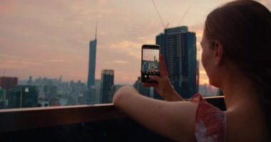 Çekici esmer kadın elinde bir telefon tutuyor ve akşam şehrinin fotoğrafını çekiyor. Terasta modern yüksek binalar var. Seyahat konsepti.