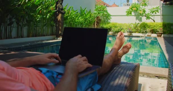 水泳プールの近くのデッキチェアに座って ラップトップで働いています 男は休暇中の遠隔作業のためにノートを使う ブロガー — ストック動画