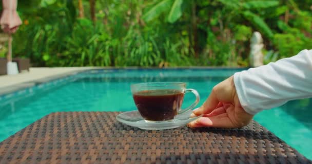 緑の熱帯林と青いスイミングプールの水と庭のソーサー上のホットコーヒーカップ 自然を背景にしたリゾートステイ バリ島 インドネシア 女の手が飲み物を持ってくる — ストック動画