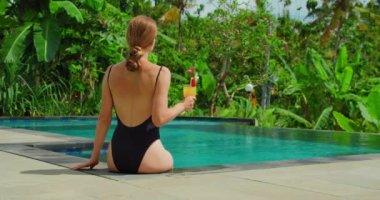 Kadının elinde bir bardak portakal suyu kokteyli ile havuz başında dinlendiği dikiz manzarası. Arka planda tropik palmiye ağaçları ve pirinç terasları var. Bali Adası Endonezya.