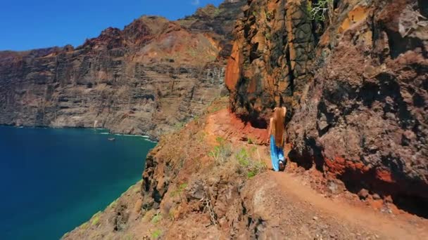 在陡峭的山坡上散步的女游客Los Gigantes 有着蓝色的深海景观 巨大的火山岩 美丽的长发姑娘爬上一座山 观察大自然的美丽 — 图库视频影像