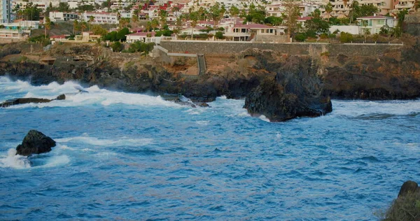 Fırtınalı okyanus ve su sıçratan dalgalar volkanik kayalara çarpıyor. Poweful mavi su manzarası. Tenerife, Kanarya Adaları, İspanya. Arka plandaki Puerto de Santiago kasabasında. Arka planda kıyıda