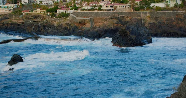 Stormy ocean and splashing waves hitting against the volcanic rocks. Poweful blue water landscape. Tenerife, Canary Islands, Spain. In the background town puerto de Santiago. In the background shore