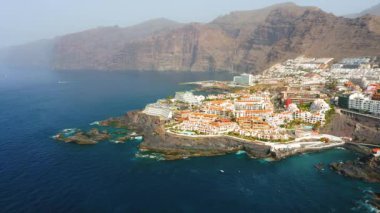Los Gigantes kayalıklarının ve siyah volkanik plajların havadan görünüşü. Otelleri, beyaz yazlıkları, binaları olan şehir manzarası. Mavi gökyüzü. Derin okyanus. Tenerife, Kanaryalar, İspanya. Muhteşem bir manzara.