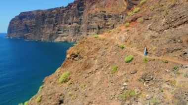 Yaz tatilinde Tenerife İspanya sahiline doğru yürüyen bir kadın. Los Gigantes kayalıkları üzerinde derin mavi gök mavisi su ve dar tehlikeli dağ yolu. Yalnız kızın dikiz manzarası.