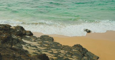 Tamamen temiz okyanus suyu, tropikal adanın vahşi sarı kumsalları ve siyah taşları. Gök mavisi turkuaz dalgalar. Sahilde farklı boyutlarda kayalar var..