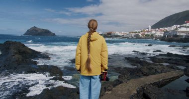 Sarı ceket ve mavi pantolon giyen esmer kız. Videograf olarak çalışan bir kadın. Atlantik Okyanusu 'nun rüzgârlı plajını çeken kadın. Ağır çekim. Tenerife. Kanarya adaları. İspanya.