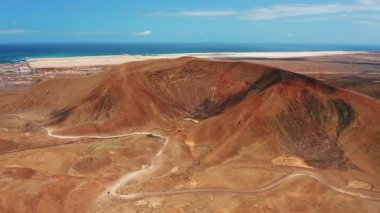 Drone flight over deserted landscape near to town Corralejo. Hiking path to volcanic empty Las Calderas, Caldera Encantada. Fuerteventura, Canary Island, Spain. Blue ocean. Cityscape. Cloudscape