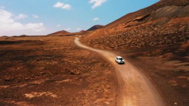 Tek başına giden beyaz bir SUV, güneşli yaz gününde çöldeki volkanik arazide boş toprak yolda seyahat ediyor. Hava aracının geniş görüşü. Araç ülke yolunda ilerliyor. Fuerteventura