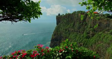 Cloudy day at Pura Luhur Uluwatu temple, Bali, Indonesia. Beautiful huge cliff with exotic plants, red magenta flowers against blue Indian Ocean. Famous historic balinese monument. Slow motion. Rain