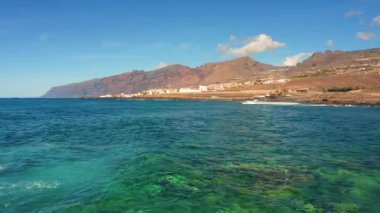 Dalgaların üzerinden uçuyor. Kayalar uzaktan görülebilir. Ufukta tatil köyü var. Turistik bir yer. Havadan deniz manzarası ve Tenerife manzarası. Deniz gelgitleri ve yukarıdan görülen gelgitler. Uçuyor