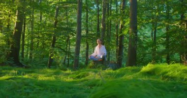 Orman. Genç Avrupalı kadın meditasyon yapıyor, yoga pozu veriyor, güneşli bahar gününde yeşil yemyeşil çayır parkı, meditasyon iyi sağlık faydaları getiriyor, spor sağlıklı yaşam tarzı getiriyor. Ağır çekim. Zihinsel