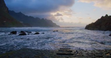 Okyanus dalgaları, Tenerife, Kanarya Adaları 'ndaki Benijo plajında kayalara çarpar ve gün batımını seyreder. Okyanus dalgalarının üzerinde gün batımı kayalık plajı yıkar. Güneş dağın tepesinde batar