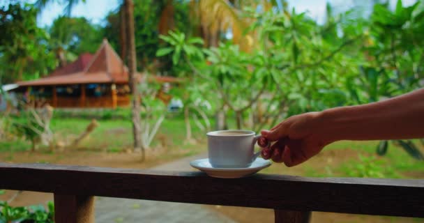 若い女性の手は 背景に美しいぼやけた自然景観とインドネシアのバリの伝統的な家とバルコニーで熱いコーヒーのカップを取ります スローモーション — ストック動画
