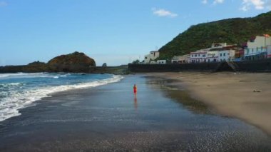 Hava görüntüsü. Sağlıklı vücut pozitifliği olan bir kadın sabah volkanik kumsalda mavi köpüklü okyanus dalgalarıyla koşuyor. Anaga Taganana kasaba dağ sırası Tenerife Kanarya Adaları İspanya. Kardiyo çalışması