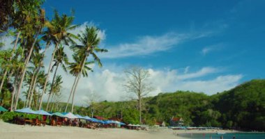 Tropik palmiye ağaçları kumlu plaj ve şeffaf sakin okyanus dalgaları. Crystal Bay Nusa Penida 'da güneşli bir günde tanınmayan insanlar. Bali Endonezya. Statik görüntü. Bulutlu mavi gökyüzü.