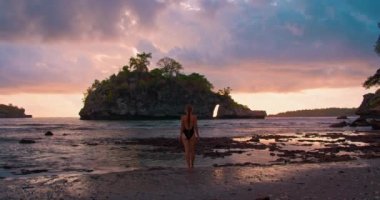 Crystal Bay Plajı 'nda güzel bir gün batımı. Nusa Penida. Arka planda mayo giymiş yalnız bir kadın okyanus dalgaları boyunca yürüyor. Turuncu ışık su yüzeyine yansıyor. Sakin deniz