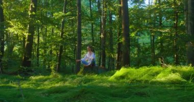 Orman. Genç Avrupalı kadın meditasyon yapıyor, yoga pozu veriyor, güneşli bahar gününde yeşil yemyeşil çayır parkı, meditasyon iyi sağlık faydaları getiriyor, spor sağlıklı yaşam tarzı getiriyor. Ağır çekim. Zihinsel