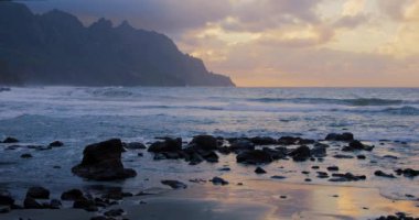 Turuncu gün doğumunda manzara kaya silueti. Okyanus dalgası kum kayalığı sahilini yavaş çekimde dağıtıyor. Deniz yüzeyi, gün batımında altın gökyüzünün ışığını yansıtıyor. Ufuk çizgisi.