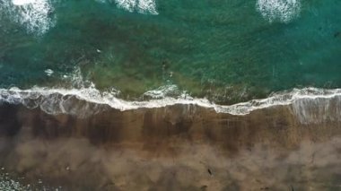 Powerful waves crash in shallow water in the Atlantic ocean. Strong currents swirl water and splash on black volcanic sand beach. Waters edge. Mesmerizing drone view.