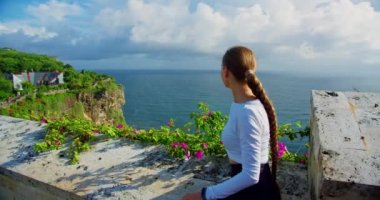 Woman using smartphone and relaxing at rocky beach. Girl taking pictures of ocean on phone. Uluwatu Bali Island Indonesia. Limestone sheer cliffs and azure Indian water. Foaming waves.