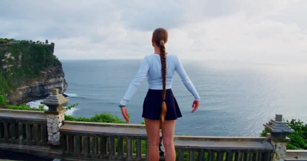 長い髪の毛を持つ女性は ウルワツ ヒンドゥー寺院プラ バリ島インドネシアで海の景色を楽しんでいます 岩の多いビーチでリラックスした観光の女の子のリアビュー 石灰岩の切り立った崖と紺碧のインド洋 — ストック動画
