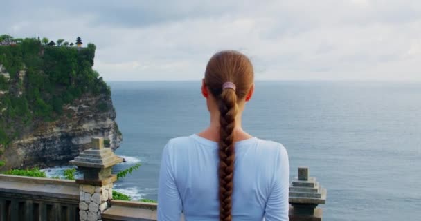 長い髪の毛を持つ女性は ウルワツ ヒンドゥー寺院プラ バリ島インドネシアで海の景色を楽しんでいます 岩の多いビーチでリラックスした観光の女の子のリアビュー 石灰岩の切り立った崖と紺碧のインド洋 — ストック動画