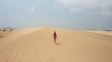 Cinematic aerial view of woman walking by sand dune. Barefoot female traveler enjoys vacations, strond wind, pink short dress. Desert empty surface with blue cloudy sky on background. Nature landscape