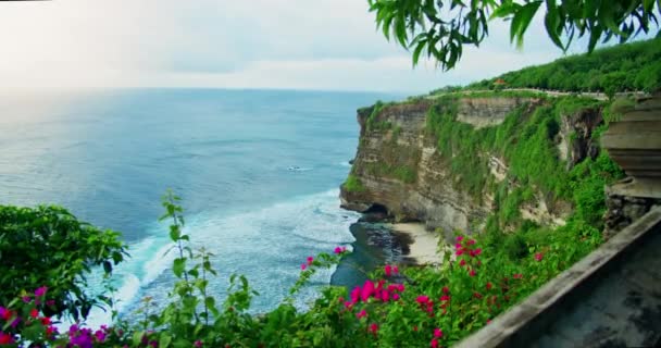 バリ島インドネシアの海を見下ろす高い崖の端に位置するウルワツ寺院 有名な観光地 熱帯の岩場の海岸に鮮やかなピンクの花を咲かせます アジア旅行 — ストック動画