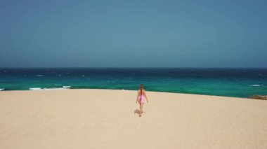 Aerial amazing view of turquoise water of ocean. Beautiful long-haired woman in dress turned away walks on golden sand dune in waterfront. Enjoyment nature. Go everywhere. Fuerteventura, Canary Island