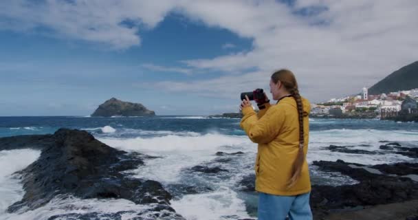 黄色のジャケットと青いパンツを着た若いブルネットの女の子 ビデオカメラマンとして働く女性 大西洋の風の強いビーチを撮影女性 スローモーション テネリフェ島 カナリア諸島 スペイン — ストック動画