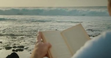 Man relaxing on beach and reading a book. Big stormy ocean waves on the background. Close-up, rear view.
