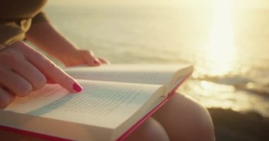 Girl self-learning by reading a book outdoors. Ocean waves on the beach at sunset. Close-up of woman hands, printed sheets of paper.
