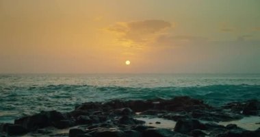 Ocean waves break on rocky shore and splashes of water fly high against sunset sky. Close-up footage.