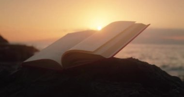 Enchanted magic open book on rocky beach at sunset glow light. Fairy tale in the pages of a book.