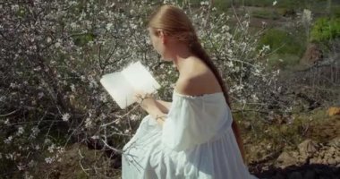 Young woman reading diary under blossoming cherry tree at backlit. Close-up of girl with open book in hands. Flowers blurred background. World poetry day.