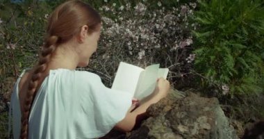 Baharın güneşli gününde çiçek açan bahçede kitap okuyan bir kadın. Beyaz elbiseli, doğal uzun örgülü saçlı bir kız..