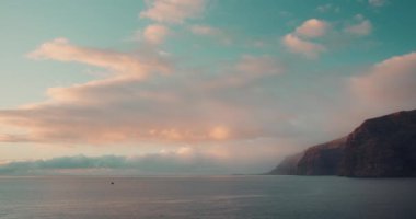 Pembe gün ışığıyla bulutlu mavi gökyüzünde güneşli yaz günbatımı Tenerife Kanarya Adaları kıyılarındaki kayalıkları ve kaya oluşumlarını aydınlatır. Los Gigantes.