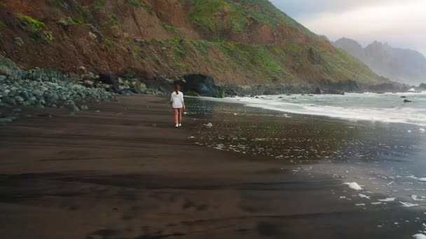 若い女性の観光客は日没時に黒い火山の黒い砂のビーチに沿って歩く テネリフェ島のカナリア島の北にあるプレヤベニョ 後ろの景色 リラックスして旅行者のコンセプトを楽しむ — ストック動画