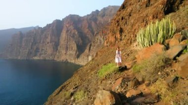 Dağın tepesinde duran beyaz elbiseli kadın okyanus manzarasının tadını çıkarıyor. Rüzgâr elbisesini üflüyor. Özgürlük, doğa kavramı. Cennet adası Tenerife, İspanya, Avrupa. Güneşli bir yaz günü. Mavi sular