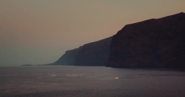 Karanlık gece okyanusunda yelken açan teknesi olan engebeli kıyı şeridi. Donanma deniz rengi, cam gibi pürüzsüz yüzeyi var. Tenerife Kanarya Adaları İspanya Avrupa. Los Gigantes uçurumları.