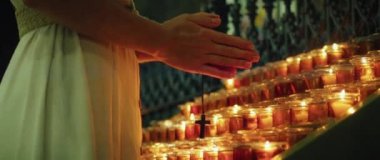 Elinde haç tutan ve tapınaktaki mumların yanında dua eden bir kadın. Yetişkin bir kız ateşli alevlerin yakınındaki günahlarını affettirmek için ellerini kavuşturdu. Doğal ateş ışığı arkaplanı.