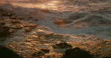 Dramatik fırtınalı okyanus dalgaları, Puerto Santiago şehrindeki kaya sahillerine ağır çekimde altın gün batımında çarpar. Deniz suyu havaya sıçrıyor ve rıhtıma yakın çekim yapıyor. Tenerife Kanaryası