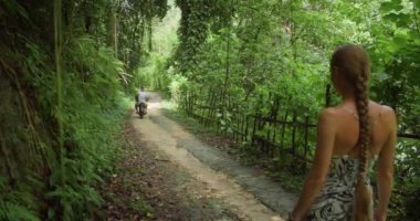Nusa Penida, Bali 'de sakin bir orman sahnesi başlıyor. Yoğun tropikal bitki örtüsüyle kaplı çamurlu bir yolda yürüyen bir kadın, motosikletli bir adamın yanından geçerken,