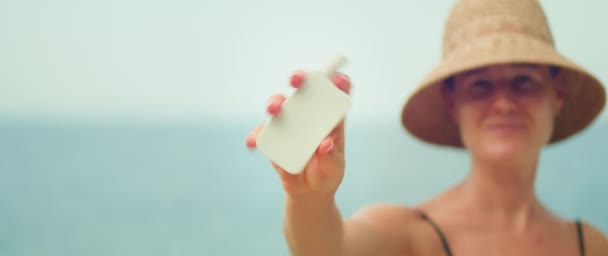 女用防晒霜管 使用前要摇匀 穿着泳衣的女孩戴着太阳帽在海滩上 护肤防晒乳霜带着大海背景 女性向相机展示化妆品 复制空间 — 图库视频影像